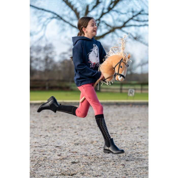 Young girl running in an outdoor riding arena wearing kids riding tights, black long riding boots, and a navy horse hoodie, holding a hobby horse.